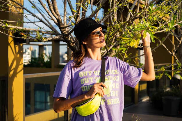 Stylish caucasian woman in trendy panama and waist neon bag on roof in Bangkok