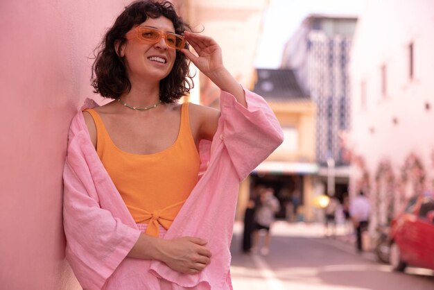Стильная кавказская молодая брюнетка в розовой летней одежде смотрит в сторону через очки, стоя на городской улице