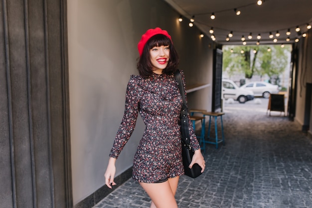 Stylish charming girl in cute vintage outfit goes to a meeting with friends after work in good mood. Portrait of young attractive brunette woman in french style clothes walking down the street