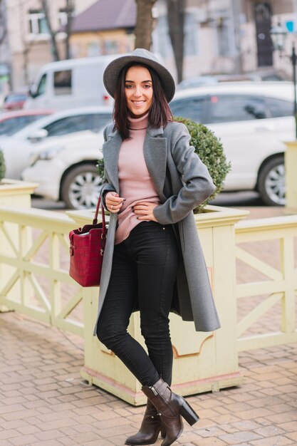 Stylish charming young woman in grey coat, hat with red bag walking on street in city centre. Brunette hair, elegant woman, fashionable model, smiling, cheerful mood.