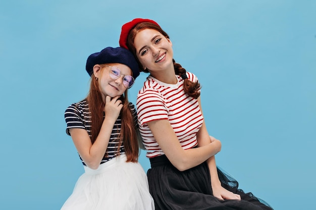 Stylish cheerful women with ginger hair in modern berets and bright clothes looking into camera on isolated blue background