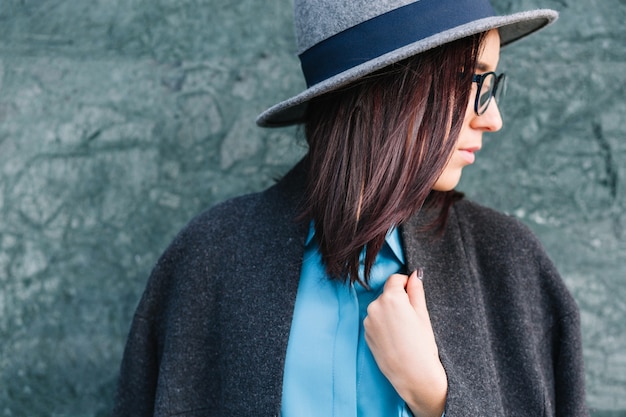 Stylish city closeup portrait fashionable young woman over grey wall. Brunette hair, black glasses, blue shirt, grey coat and hat. Elegant outlook, true emotions, looking to side.