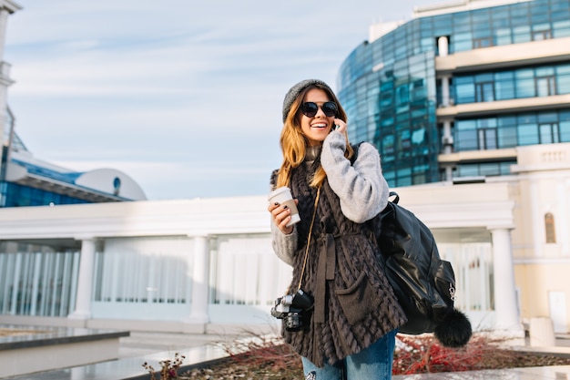 Стильный городской портрет модной красивой девушки, идущей с кофе в центре современного города Европы. Радостная молодая женщина в зимнем теплом свитере