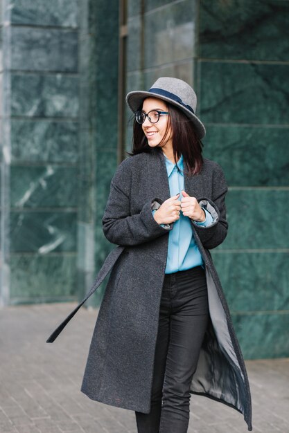 세련 된 도시 초상화 세련 된 젊은 여자 거리에 긴 회색 코트에 산책. 모자, 검은 안경을 쓰고, 측면에 웃고, 진정한 행복 감정을 보여주는 젊은 사업가.