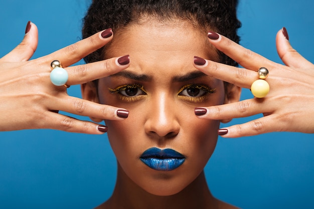 Стильная концентрированная афро женщина с ярким макияжем демонстрирует кольца на пальцах, держа руки на лице, изолированном над синей стеной
