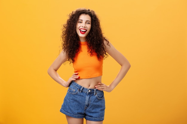 Elegante donna sicura e fantastica pronta alla festa accesa in piedi in top corto alla moda e pantaloncini di jeans con le mani in vita che sporgono la lingua e sorride con gioia in posa su sfondo arancione.