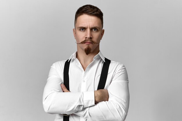 Stylish confident bearded boss in suspenders and white shirt posing isolated at blank copyspace studio wall