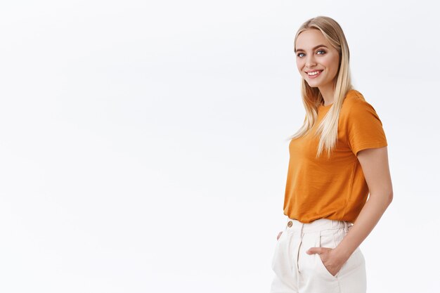 Stylish, confident good-looking female coworker with blond hair, wear orange t-shirt, half-turned look camera self-assured, hold hands in pants, smiling like professional, white background