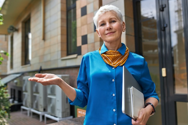 Stylish confident middle aged businesswoman with short hairstyle posing outside office building with laptop under her arm, making gesture as if holding something on hand