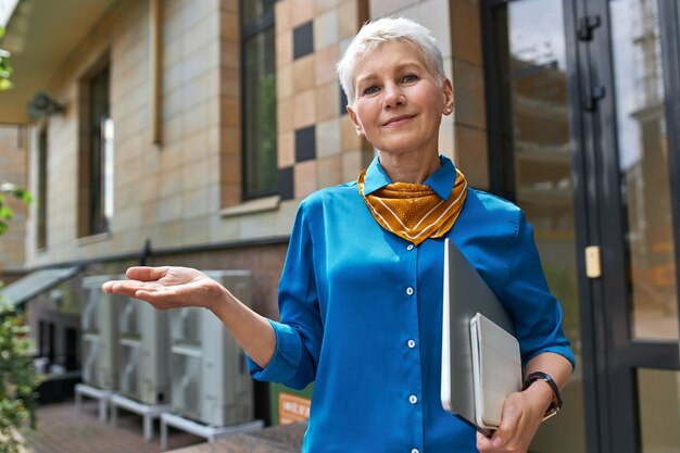 Stylish confident middle aged businesswoman with short hairstyle posing outside office building with laptop under her arm, making gesture as if holding something on hand