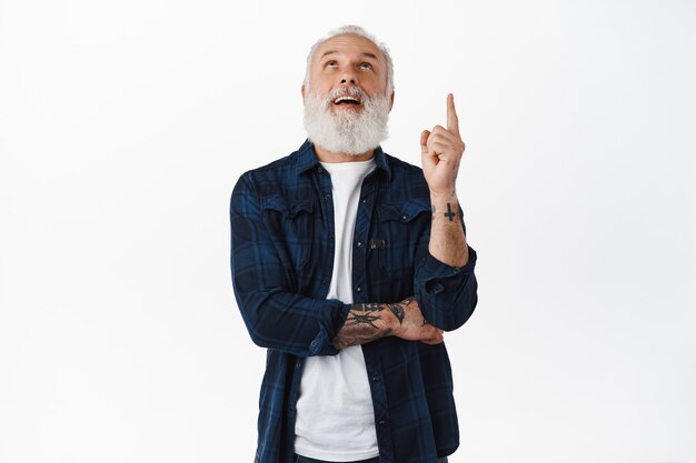 Stylish cool grandfather with tattoos and long beard, looking and pointing finger up at advertisement, staring above at promotional text copyspace, standing over white wall