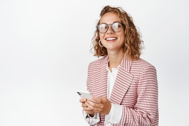 Stylish corporate woman in suit and glasses, holding mobile phone and smiling on white