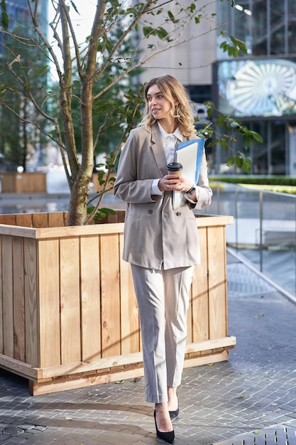 Stylish corporate woman in suit standing on street with documents work laptop and coffee waiting nea