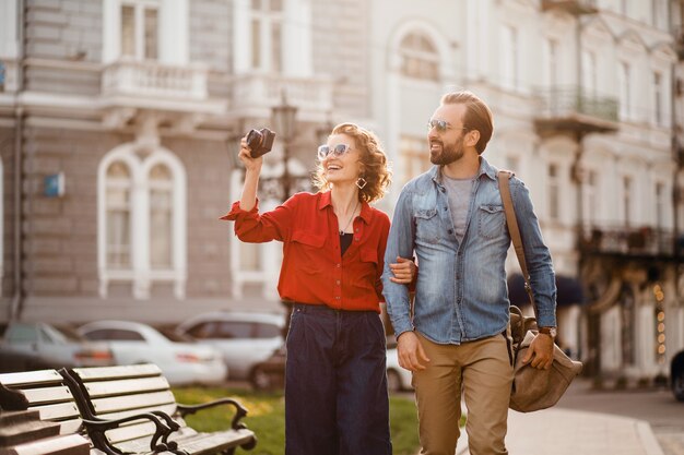 Stylish couple in love walking embracing in street on romantic trip and taking photo