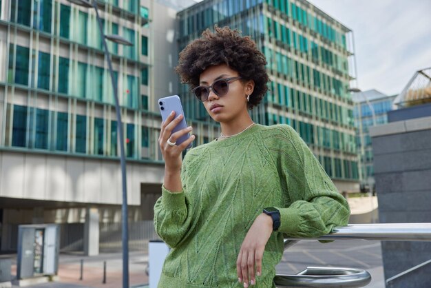スタイリッシュな縮れ毛の女性はサングラスをかけ、緑のジャンパーはスマートフォンを使用して、フォロワーとのストリートチャットで携帯電話アプリケーションを介して都市の設定検索場所に対してメッセージポーズを取得します