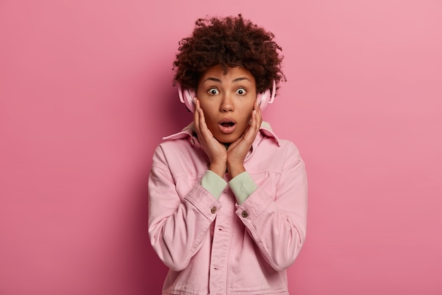 Stylish curly haired young woman keeps hands on cheeks, wears pink jacket, listens radio online, uses wireless headphones, has shocked face expression