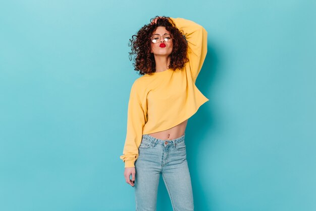 Stylish curly woman with red lipstick touches hair and blows kiss. Portrait of girl in glasses and yellow sweater on blue space.