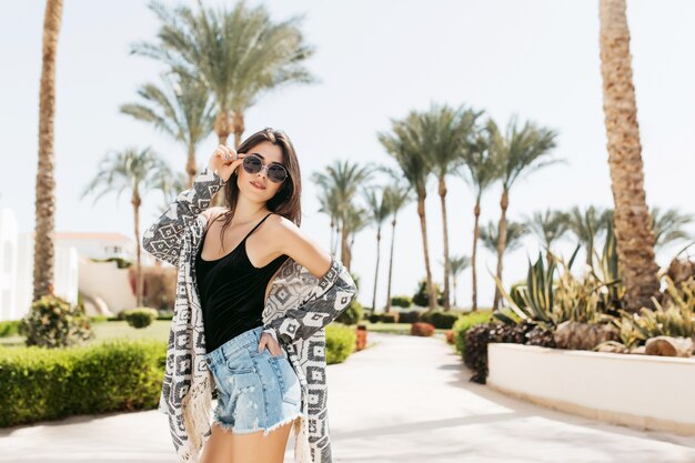 Stylish cute girl in black tank-top and denim shorts holding her sunglasses while posing with palm trees. Charming young woman in trendy attire having fun on the luxury resort in summer