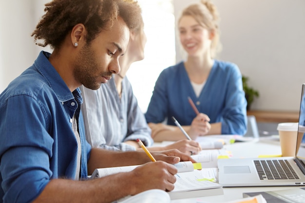 Elegante uomo dalla pelle scura in camicia blu, impegnato con lo studio, seduto vicino ai suoi compagni di gruppo femminili, computer portatile funzionante, scrittura di carta di diploma. gruppo di studenti amichevoli di razze diverse