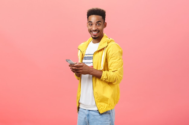 Stylish delighted african american male with afro curly haircut standing half-turned over pink wall holding smartphone wearing yellow trendy jacket smiling joyfully showing friend features of device.