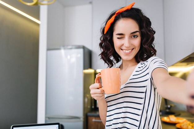 笑みを浮かべて、お茶を楽しんでかなり若い女性のスタイリッシュな国内selfieの肖像画。自宅での週末、モダンなアパートメント、レジャー、心地よさ