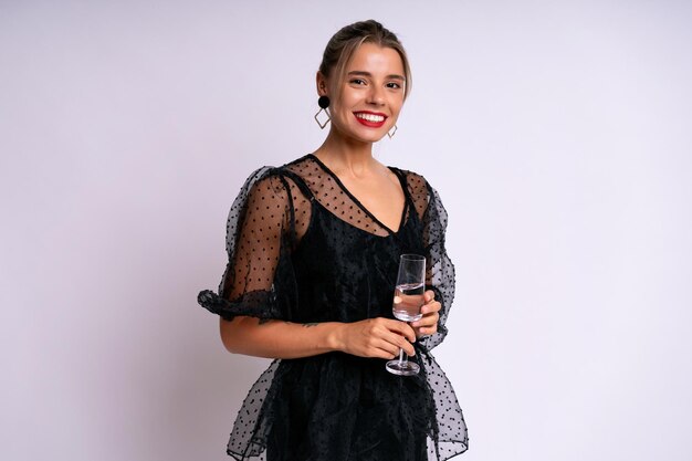 Стильная элегантная женщина в черном вечернем платье, держащая стакан с напитком, готовая к празднованию, на белом фоне.