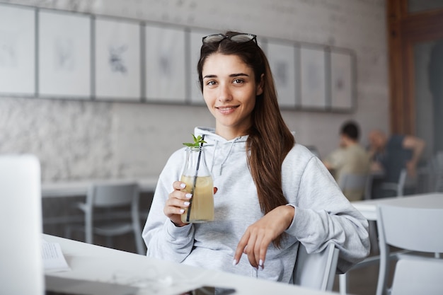 Elegante ed elegante giovane studentessa in un caffè, bevendo cocktail e sorridendo ampiamente, mentre si prende una pausa dal lavoro che fa tramite computer portatile.