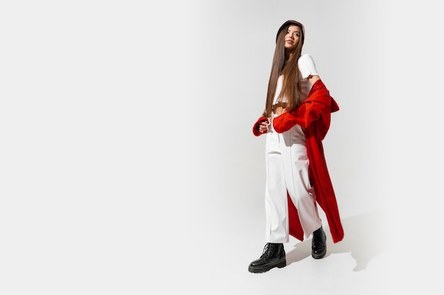 Stylish european brunette woman in red coat and black hat posing on white wall