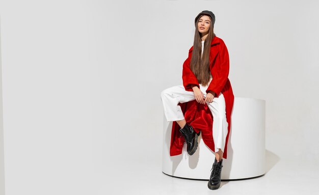 Stylish european brunette woman in red coat and black hat posing on white wall