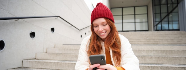 Free photo stylish european girl with red hair sits on public stairs with smartphone places online order sends