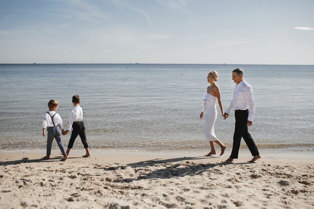 Stylish family is walking on the beach near the calm sea, parents and children are holding hands together