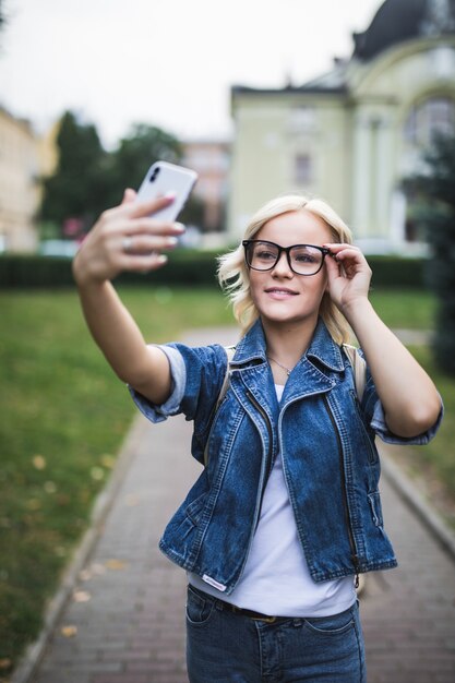 Stylish fashion blonde girl woman in jeans suite and glasses makes selfie on her phone in the city in the morning