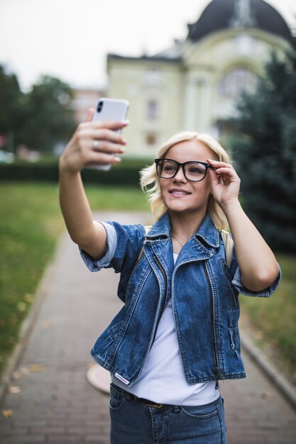 청바지 스위트와 안경에 세련된 패션 금발 소녀 여자는 아침에 도시에서 그녀의 전화에 셀카를 만든다