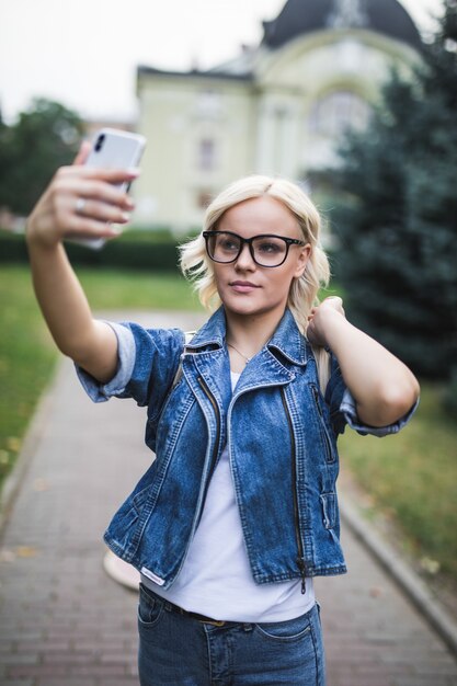 ジーンズスイートのスタイリッシュなファッションのブロンドの女の子の女性は、午前中に街で彼女の電話でselfieを作る