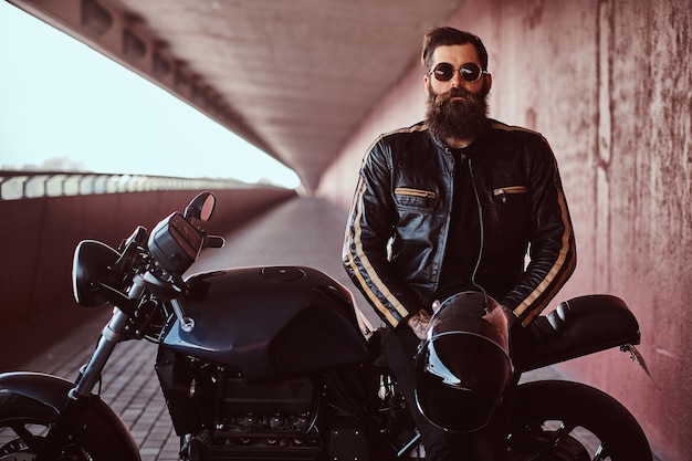 Stylish fashionable biker dressed in a black leather jacket with sunglasses holds a helmet sitting on his custom-made retro motorcycle, looking at a camera.