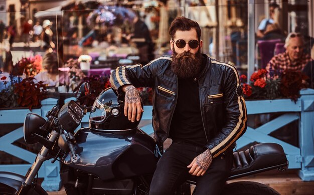カフェのテラスの近くで彼のカスタムメイドのレトロなオートバイに座って、黒い革のジャケットを着たサングラスをかけたスタイリッシュでファッショナブルなバイカー。