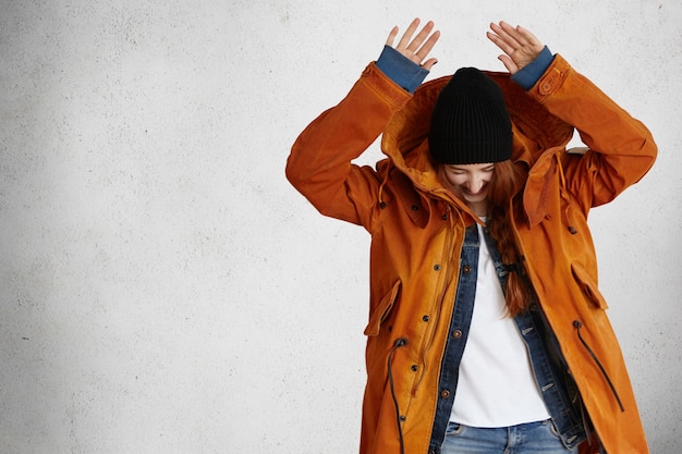 Stylish and fashionable redhead model in red winter coat and trendy black hat keeping her hands above head