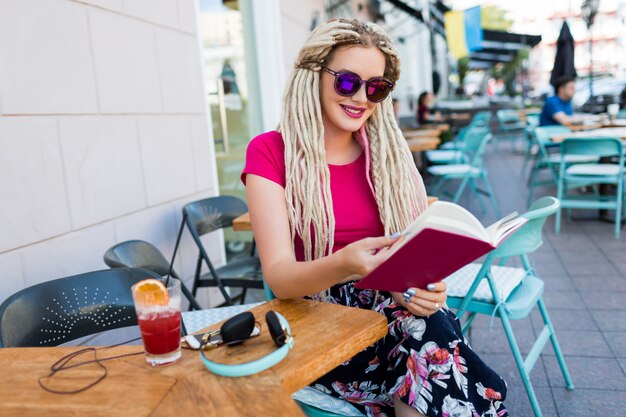 Стильная модная женщина с белыми дредами в солнцезащитных очках держит ноутбук и проводит свободное время в современном ресторане. Свежий коктейль и наушники на столе.