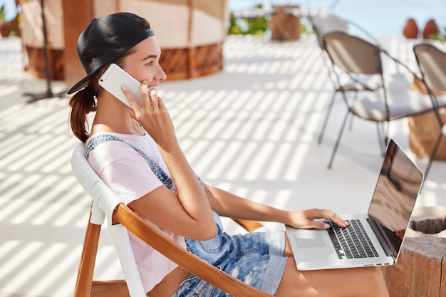黒い帽子のスタイリッシュな女性、カジュアルな白いtシャツ、ラップトップコンピューターのオンラインチャットのメッセージ、スマートフォンの親友との会話