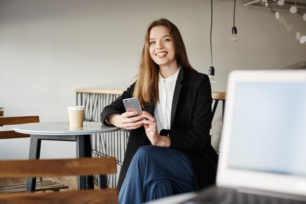 携帯電話でカフェに座っているスタイリッシュな女性起業家、笑みを浮かべて