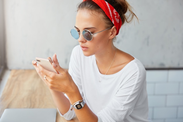 Stylish female student wears sunglasses, red headband and white sweater, downloads files on mobile phone, sits in coffee shop, has serious expression. Woman uses modern technologies and internet