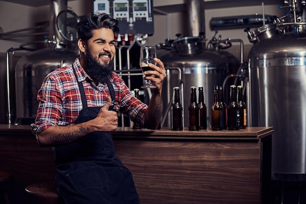 Foto gratuita elegante uomo indiano con la barba completa in una camicia in pile e grembiule tiene un bicchiere di birra, seduto dietro il bancone in una fabbrica di birra.