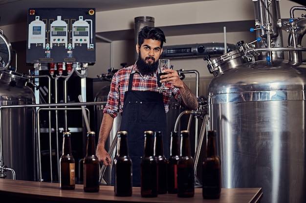 Стильный бородатый индийский мужчина в флисовой рубашке и фартуке держит стакан пива, стоя за прилавком пивоварни.