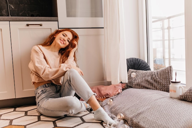 Stylish ginger girl relaxing at home Redhaired laughing woman in jeans sitting on the floor