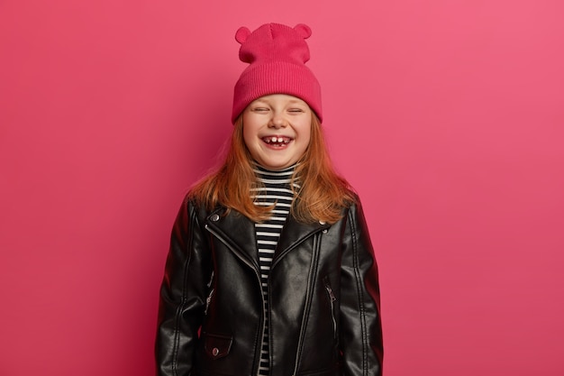 Стильная рыжая милая девочка искренне смеется, закрывает глаза, очень рада, радуется хорошему дню, проводит свободное время с мамой и папой, носит розовую шляпу и кожаную куртку, готовится к школе