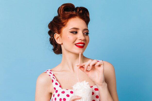 Stylish ginger woman enjoying milkshake on blue space. Smiling caucasian girl drinking beverage.