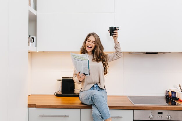 Stylish girl in blue jeans raising cup of tea and laughing