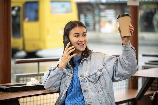 Stylish girl in casual style speaks on the phone with coffee in hand and is waiting for someone.