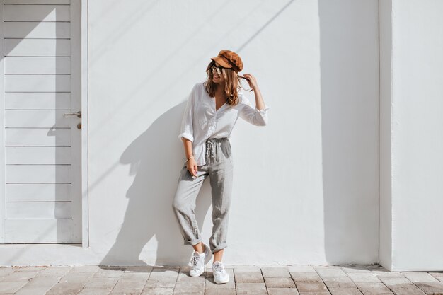 Stylish girl in gray pants and white cotton blouse posing near white wall. woman in cap and glasses.