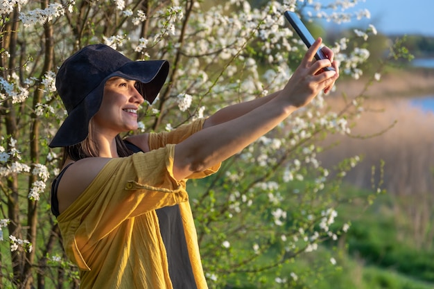 A stylish girl in a hat makes a selfie at sunset near flowering trees in the forest.